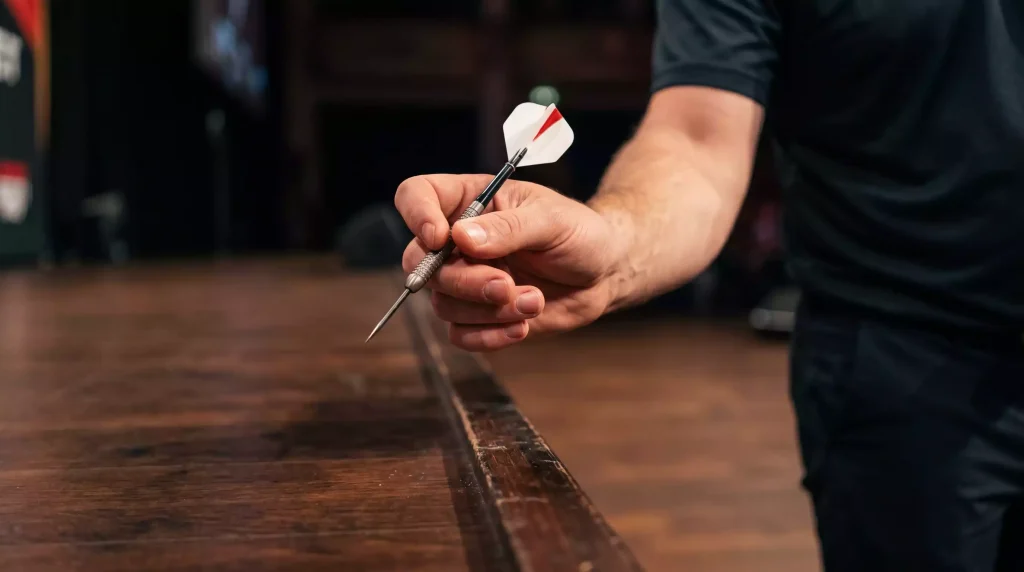 Detailaufnahme der Hand eines Profi-Dartspielers im präzisen Drei-Finger-Griff an einem Tungsten-Steel-Tip-Pfeil, im unscharfen Hintergrund die markierte Oche-Linie auf dem Turnierboden.