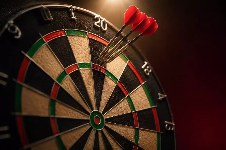 Drei Tungsten-Steel-Tip-Pfeile dicht beieinander in der Triple-20-Zone einer Sisal-Dartscheibe, warmes Bühnenlicht von oben, tiefdunkler Hintergrund.